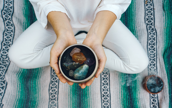 A person in a cross-legged position holding a singing bowl filled with various polished tumble stones like amethyst and rose quartz